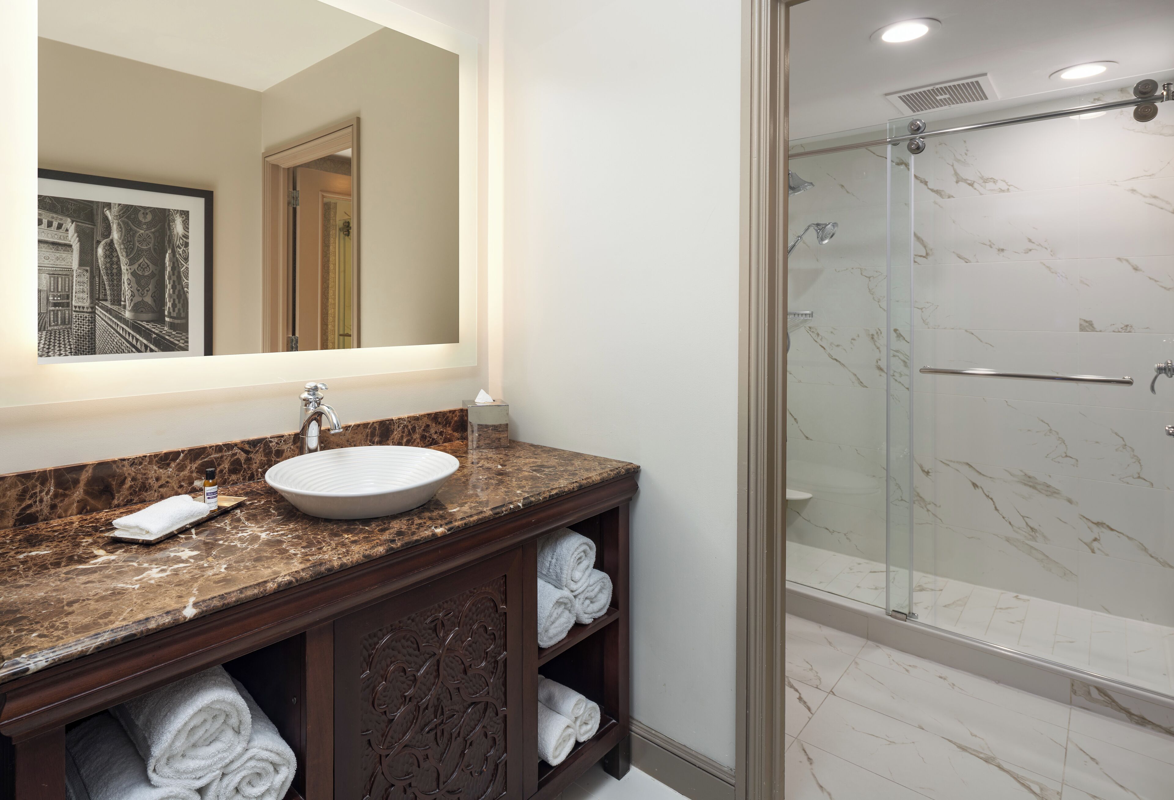 A bathroom with a marble countertop, a sink, a glass shower enclosure, and a mirror with a framed picture.