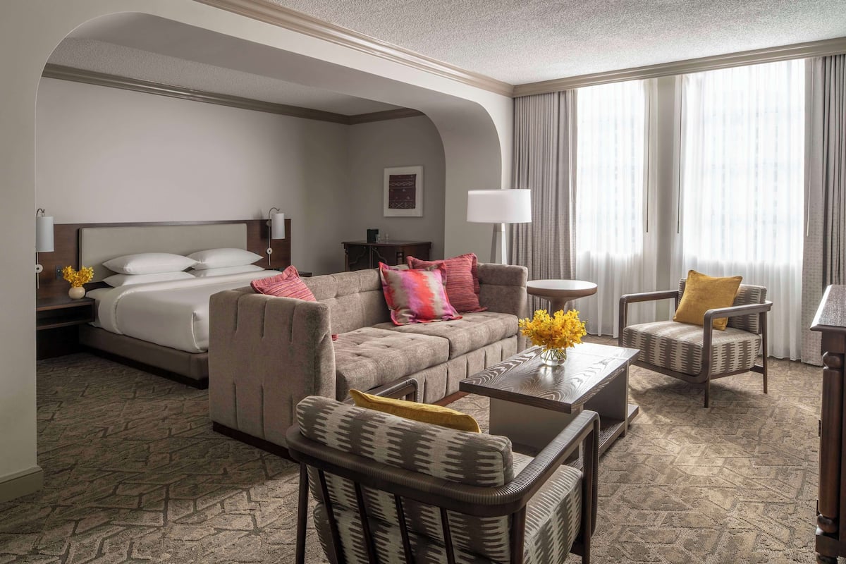 A hotel room with a bed, a sofa, a chair, a small table, and a lamp.