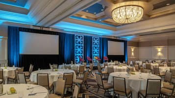 A banquet hall with round tables, chairs, a stage with a screen, and a chandelier.