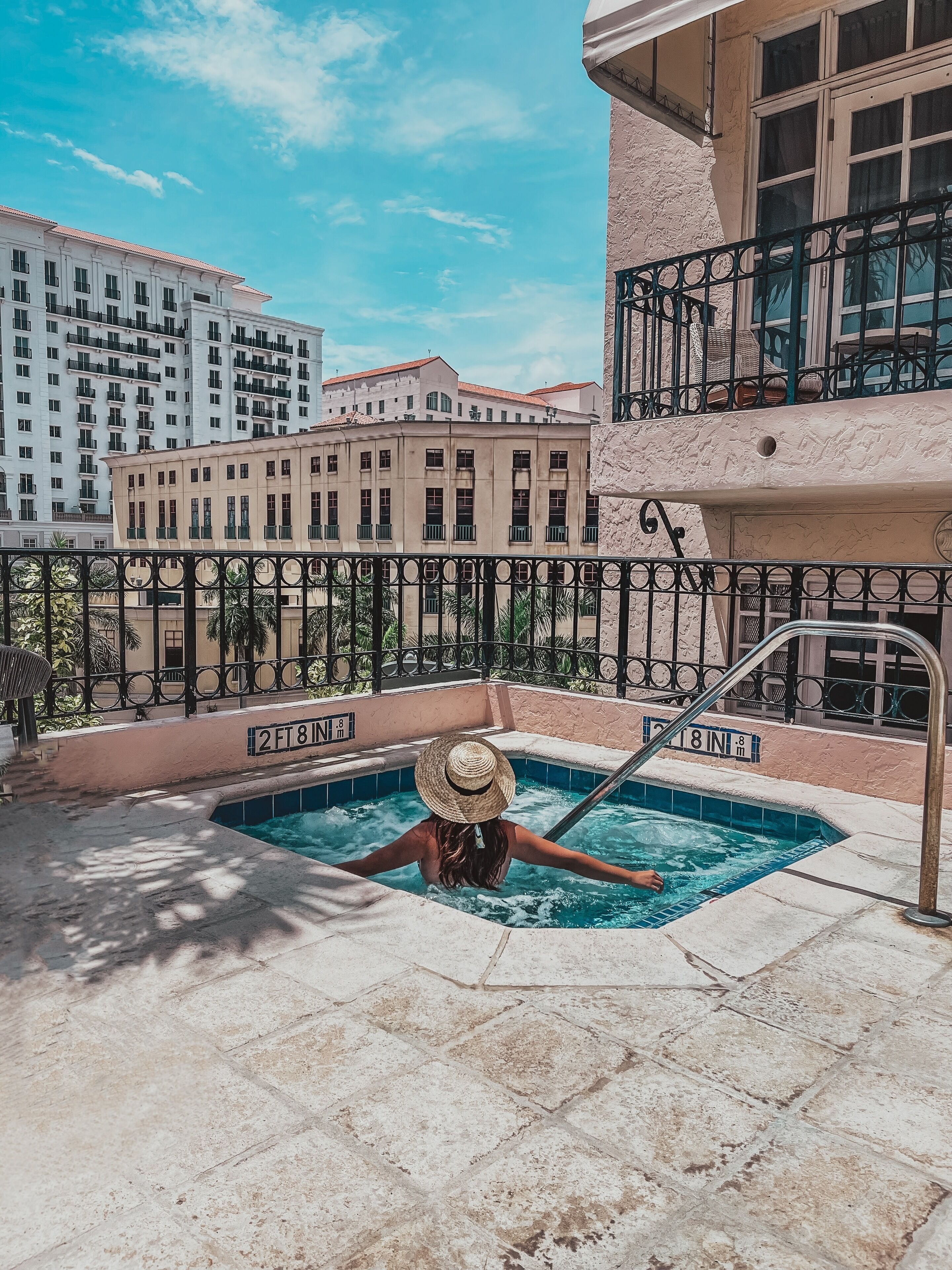 A person in a hot tub on a balcony with a view of buildings and a clear sky.