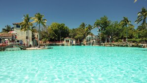 Une grande piscine d’un bleu turquoise éclatant, entourée de palmiers et de bâtiments, avec un pont enjambant l’eau.