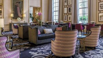 Un vestíbulo de hotel con un sofá, sillones estampados, una zona de comedor y ventanas amplias.