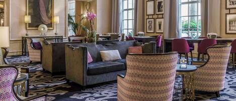Un vestíbulo de hotel con un sofá, sillones estampados, una zona de comedor y ventanas amplias.