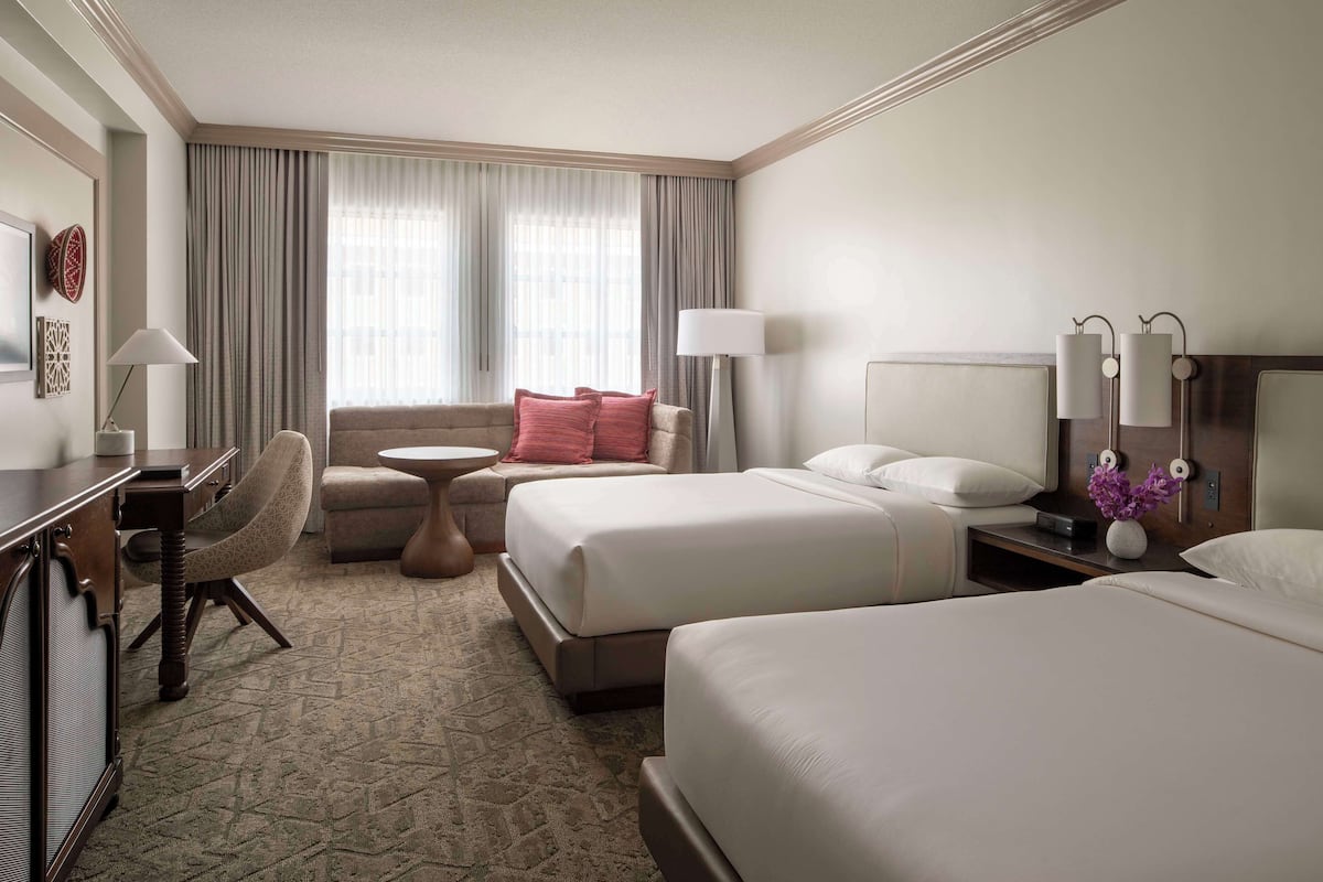 A hotel room with two beds, a desk, a chair, a small table, and a lamp.
