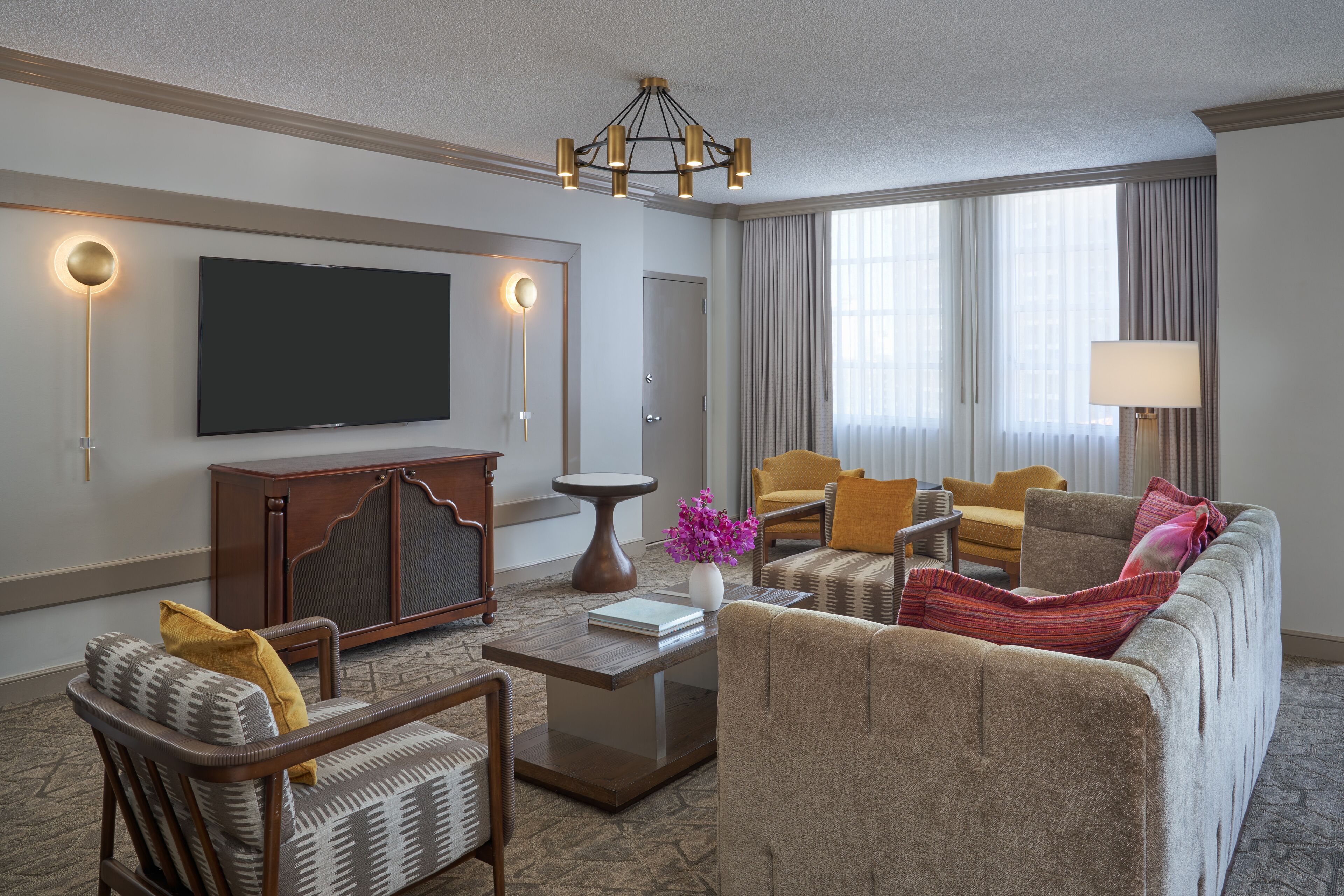 Una habitación de hotel moderna con televisión de pantalla plana, un mueble de madera, una mesa de centro redonda y un sofá beige con cojines de colores.