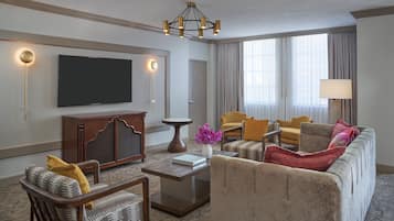 Una habitación de hotel moderna con televisión de pantalla plana, un mueble de madera, una mesa de centro redonda y un sofá beige con cojines de colores.