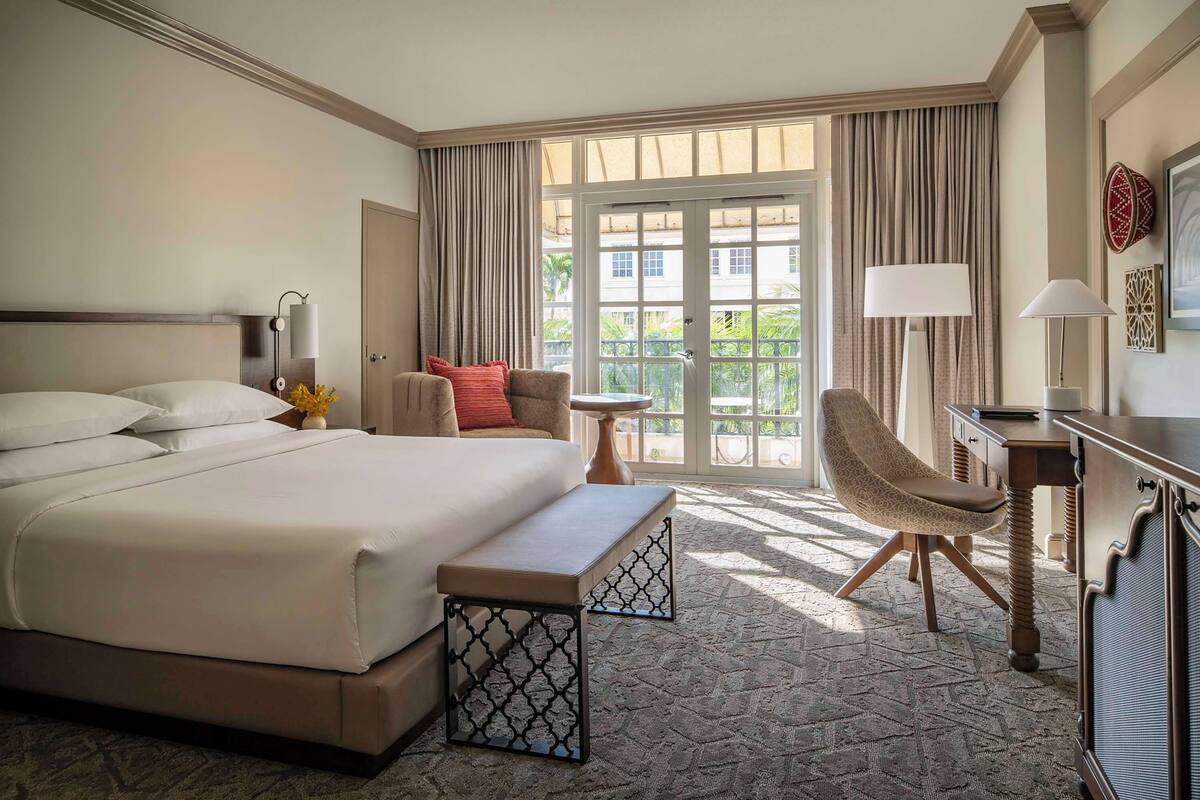 A hotel room with a large bed, a desk, a chair, and a balcony with curtains.