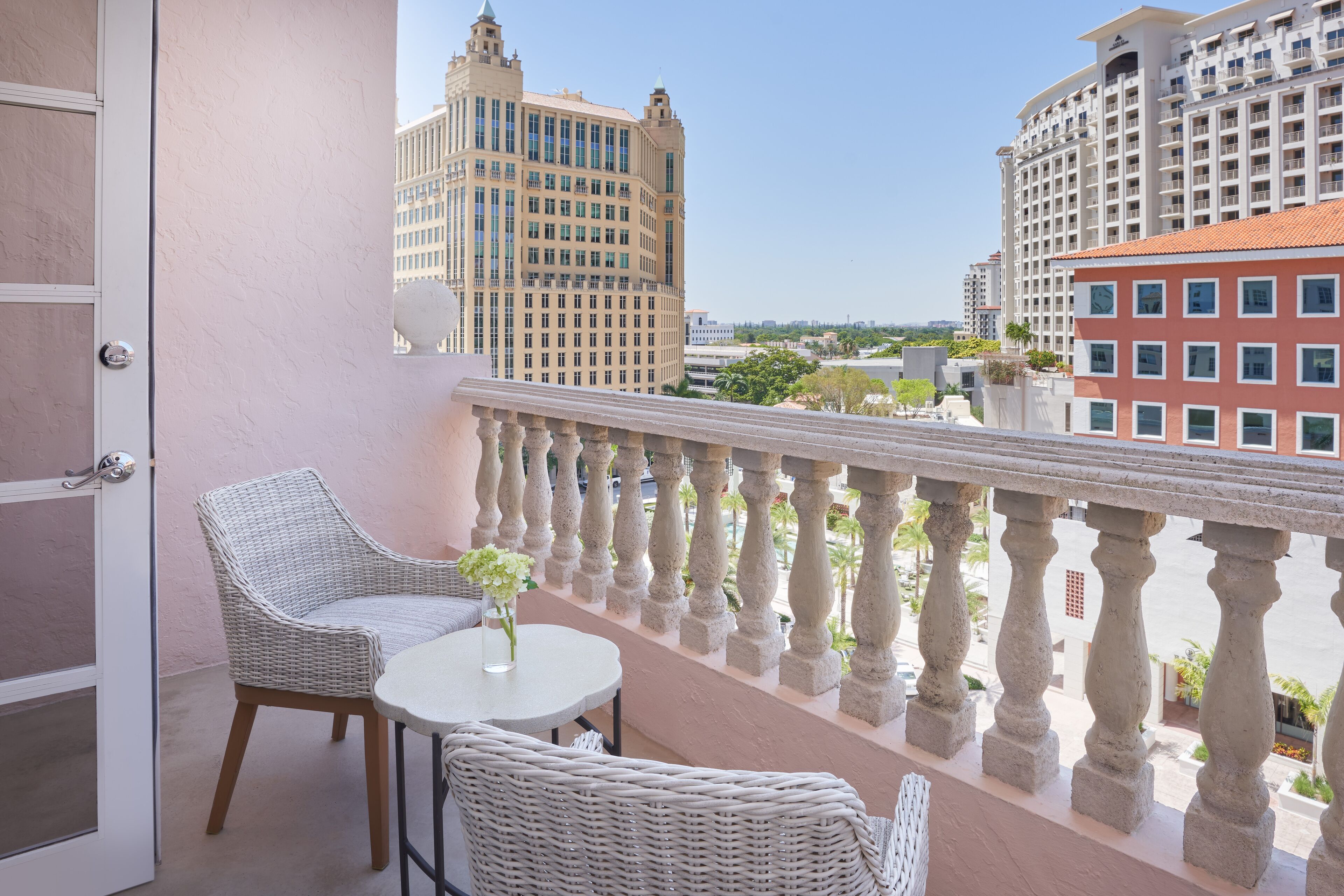 A balcony with wicker furniture and a small table, overlooking a cityscape with tall buildings.
