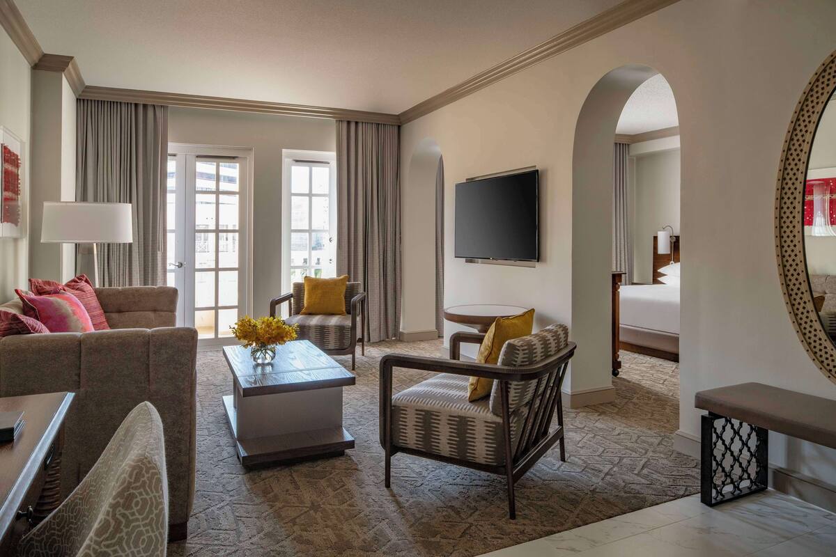 A hotel room with a sofa, armchair, coffee table, and a TV. There is a bed with a large mirror and a window with curtains.