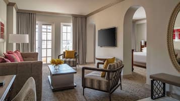 Ein Hotelzimmer mit Sofa, Sessel, Couchtisch und Fernseher. Es gibt ein Bett mit großem Spiegel und ein Fenster mit Vorhängen.