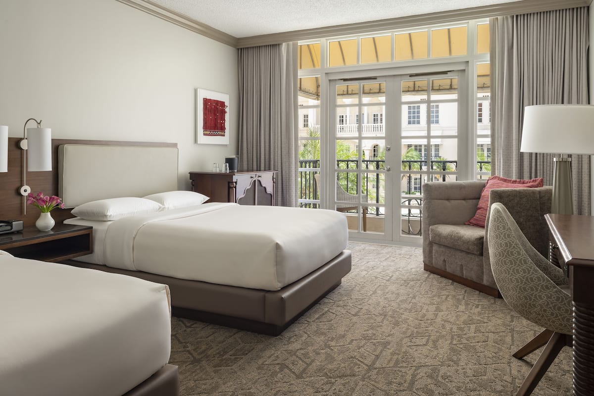 A hotel room with a large bed, a desk, a chair, and a balcony with a view of buildings.