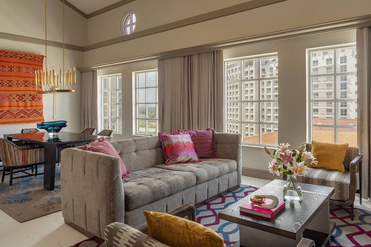 A modern living room with a sofa, coffee table, and chairs. There is a large window with a view of buildings.