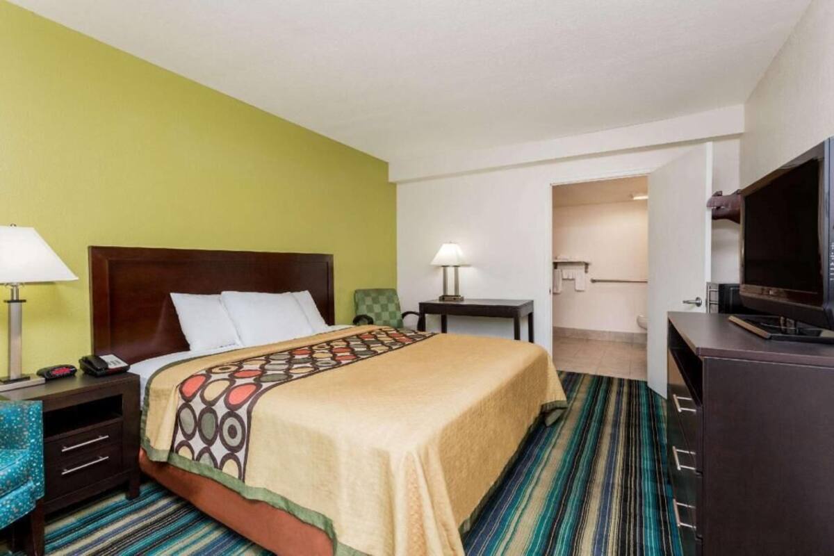 A hotel room with a bed, bedside table, lamp, TV, and a view of the hallway.