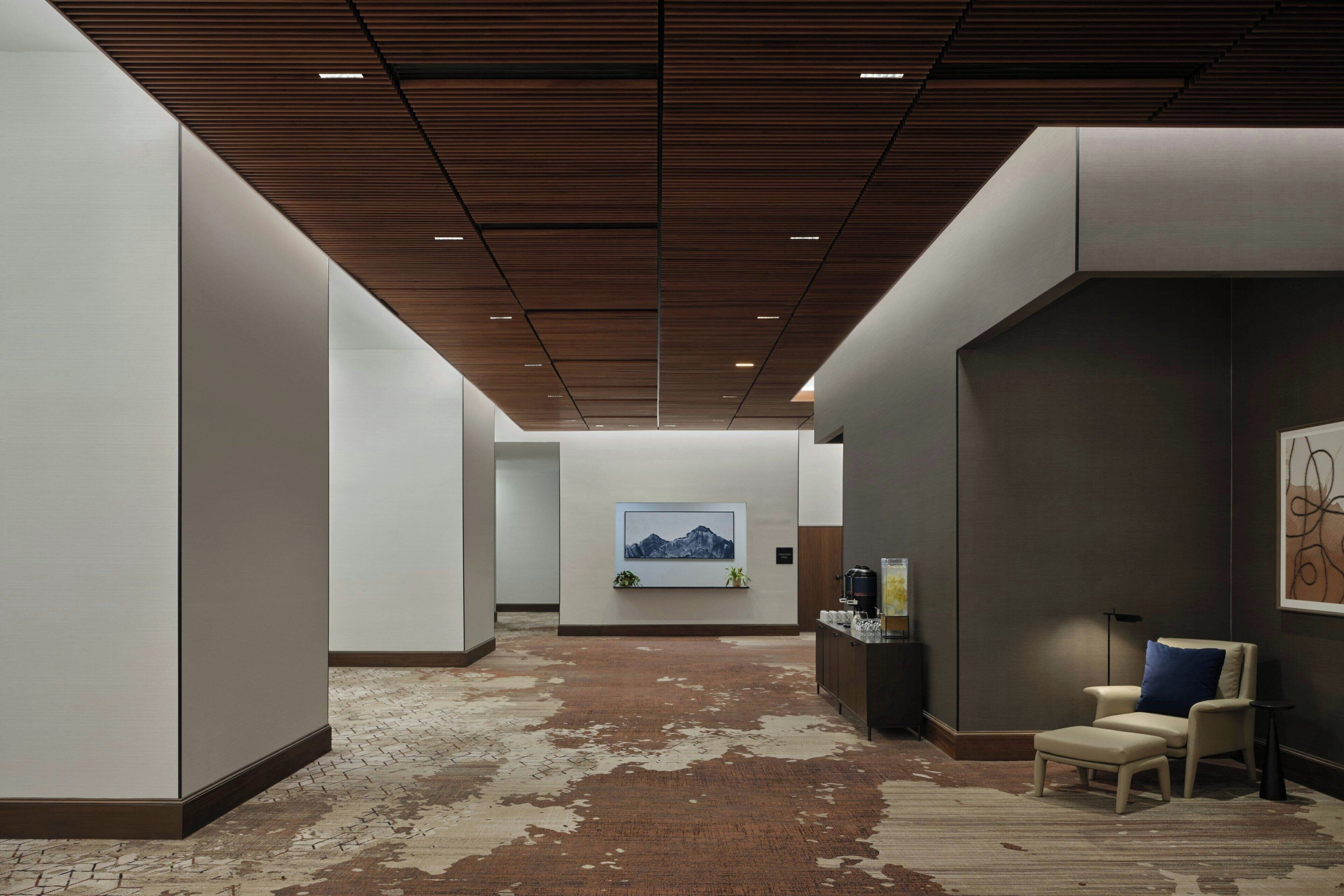 Un corridoio moderno con soffitto in legno, un quadro appeso, un’area salotto con una poltrona e poggiapiedi, e un tavolino con una lampada.