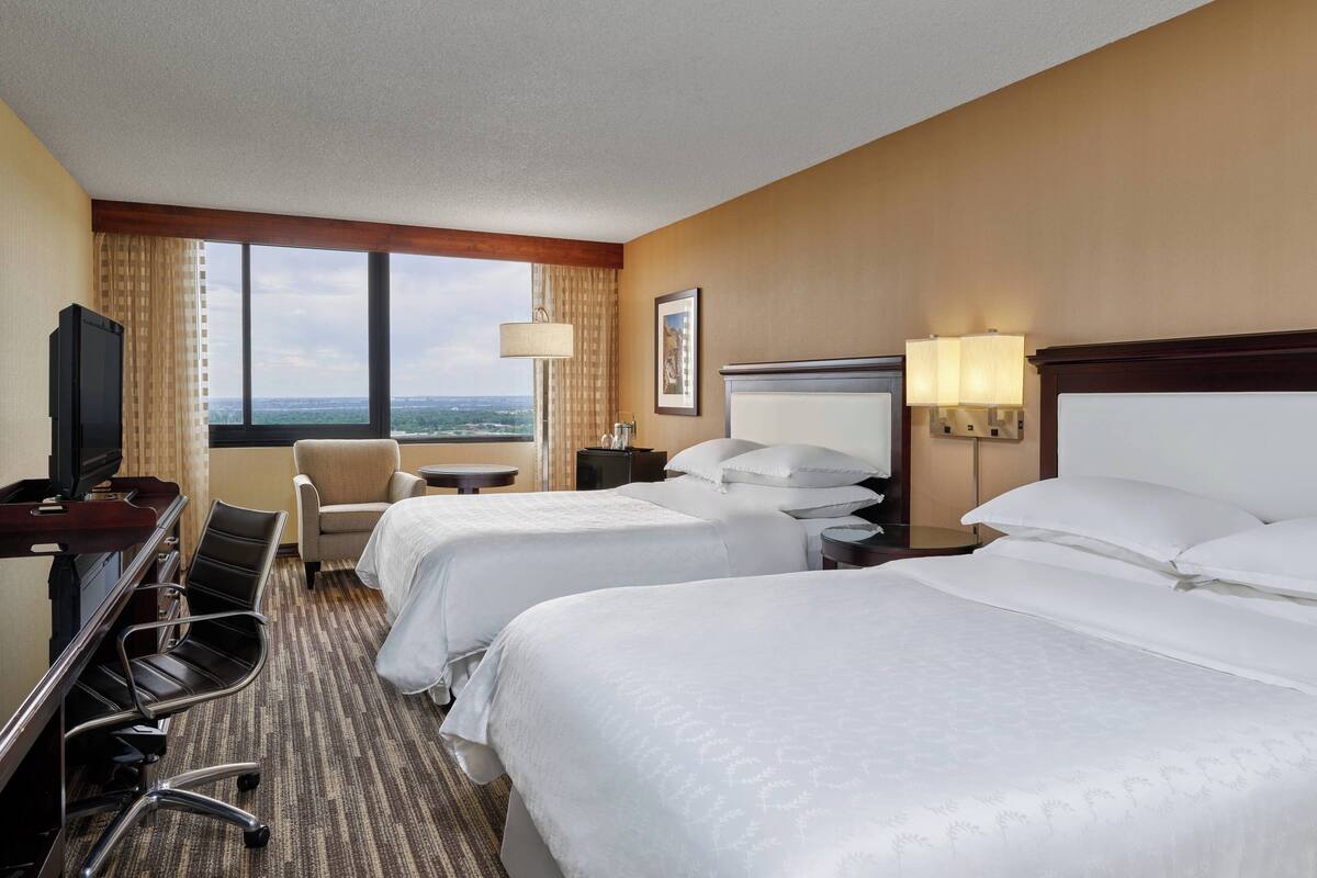 A hotel room with two beds, a desk with a chair, a television, and a view of the city.