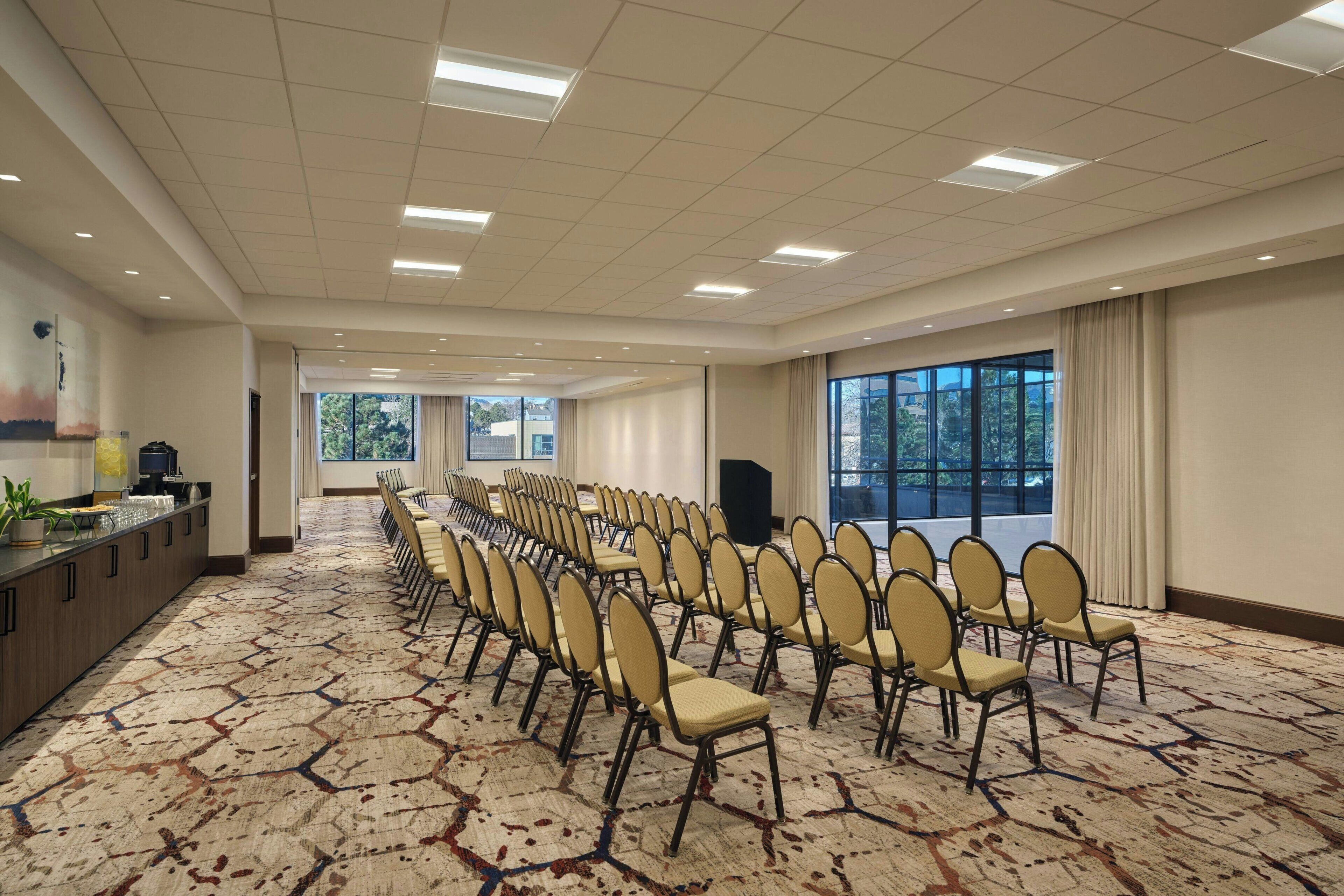 Un salón de conferencias con filas de sillas, una mesa larga y ventanas amplias.