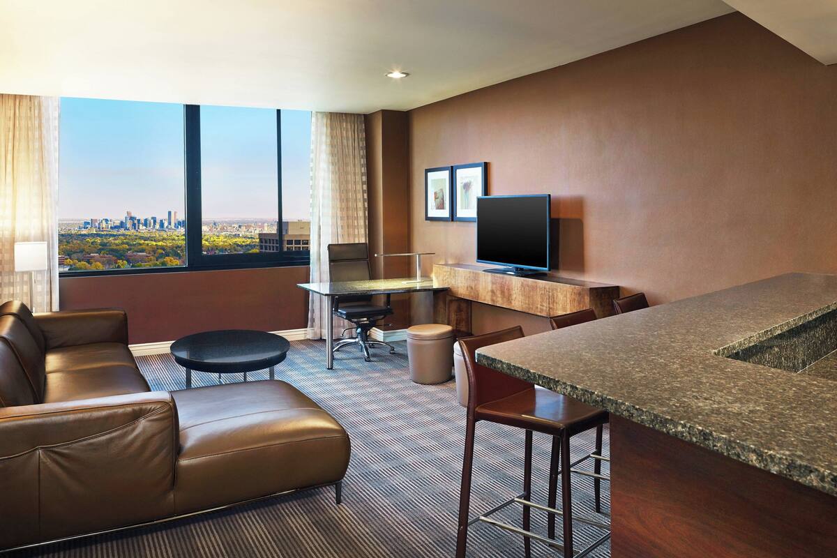A modern hotel room with a large window offering a city view, a leather sofa, a small round table, a TV, and a bar area.