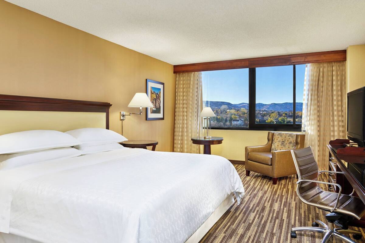 A hotel room with a large bed, a desk with a chair, a television, and a window with a mountain view.
