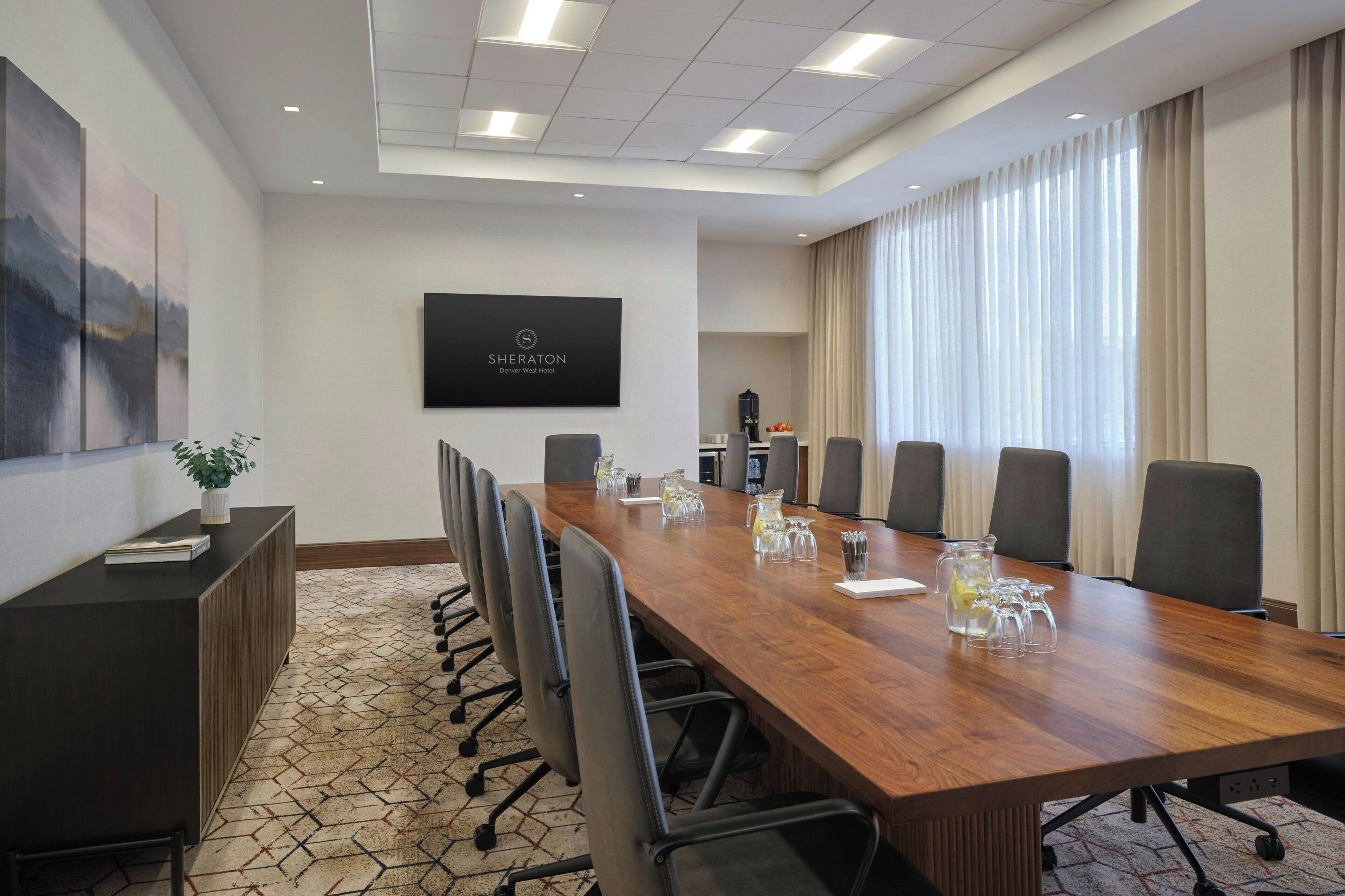 Uma sala de conferências com uma longa mesa de madeira, várias cadeiras, uma tela preta com a marca do hotel e uma grande janela com cortinas.