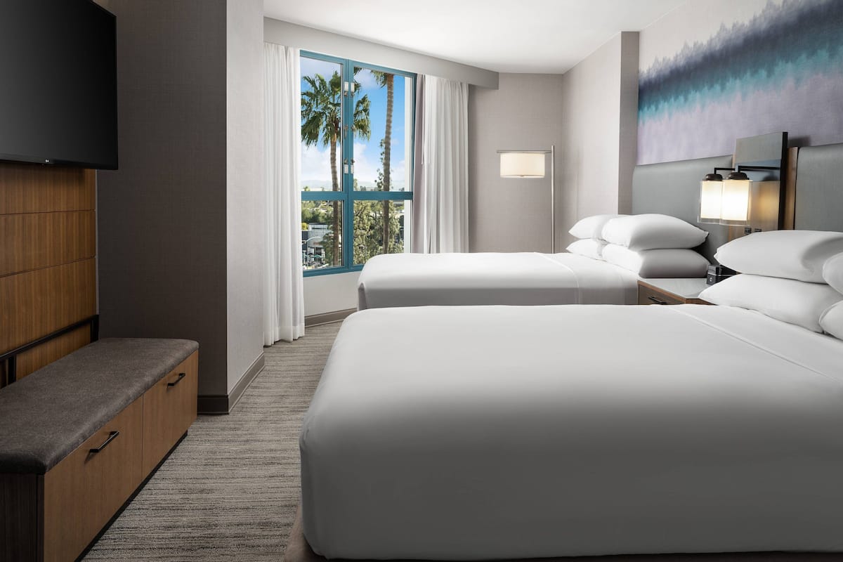 Habitación de hotel con dos camas, televisor, escritorio y ventana con vista a palmeras.