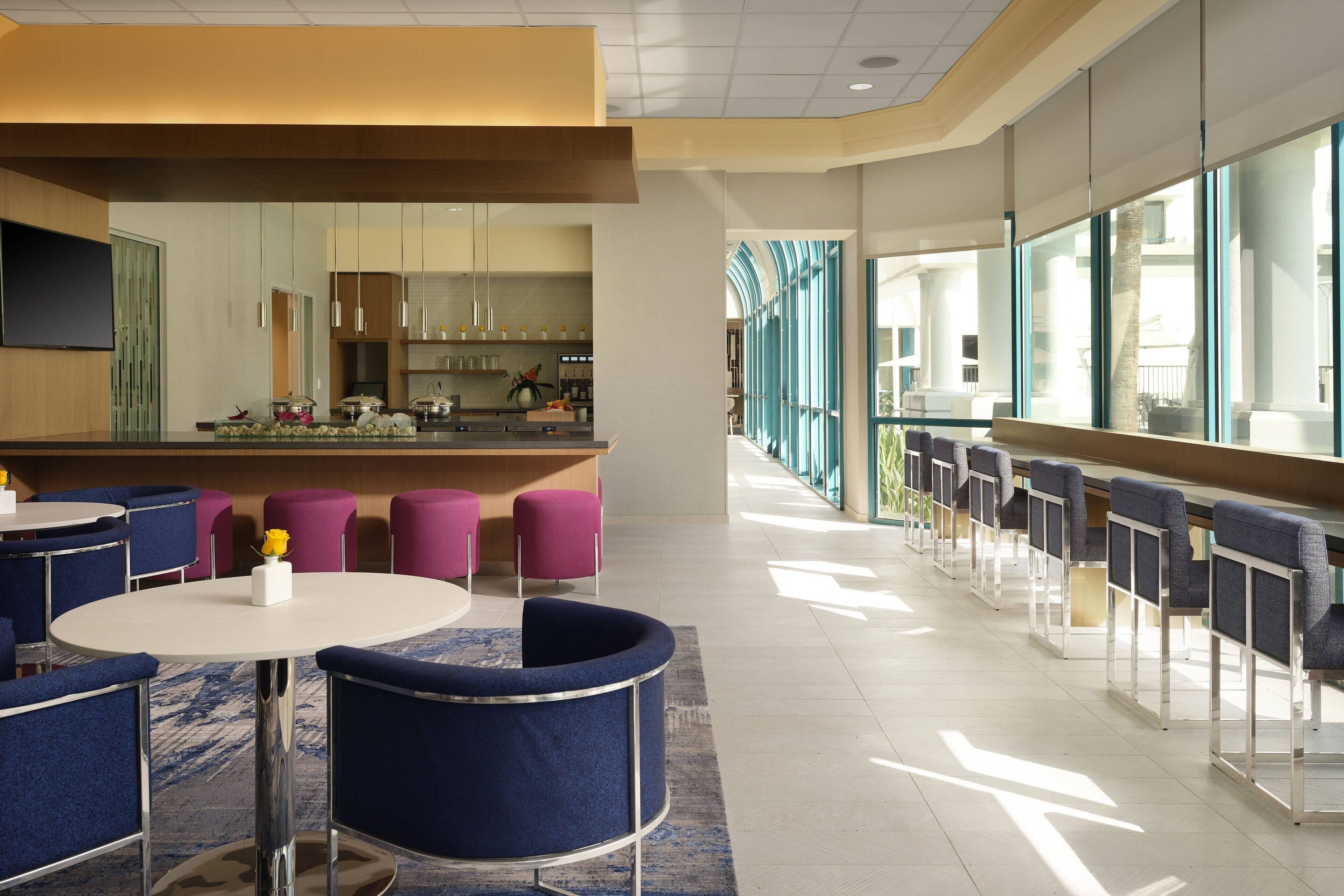 Eine moderne Hotellobby mit runden Tischen und blauen Sesseln, einer Bar und großen Fenstern mit Blick ins Freie.