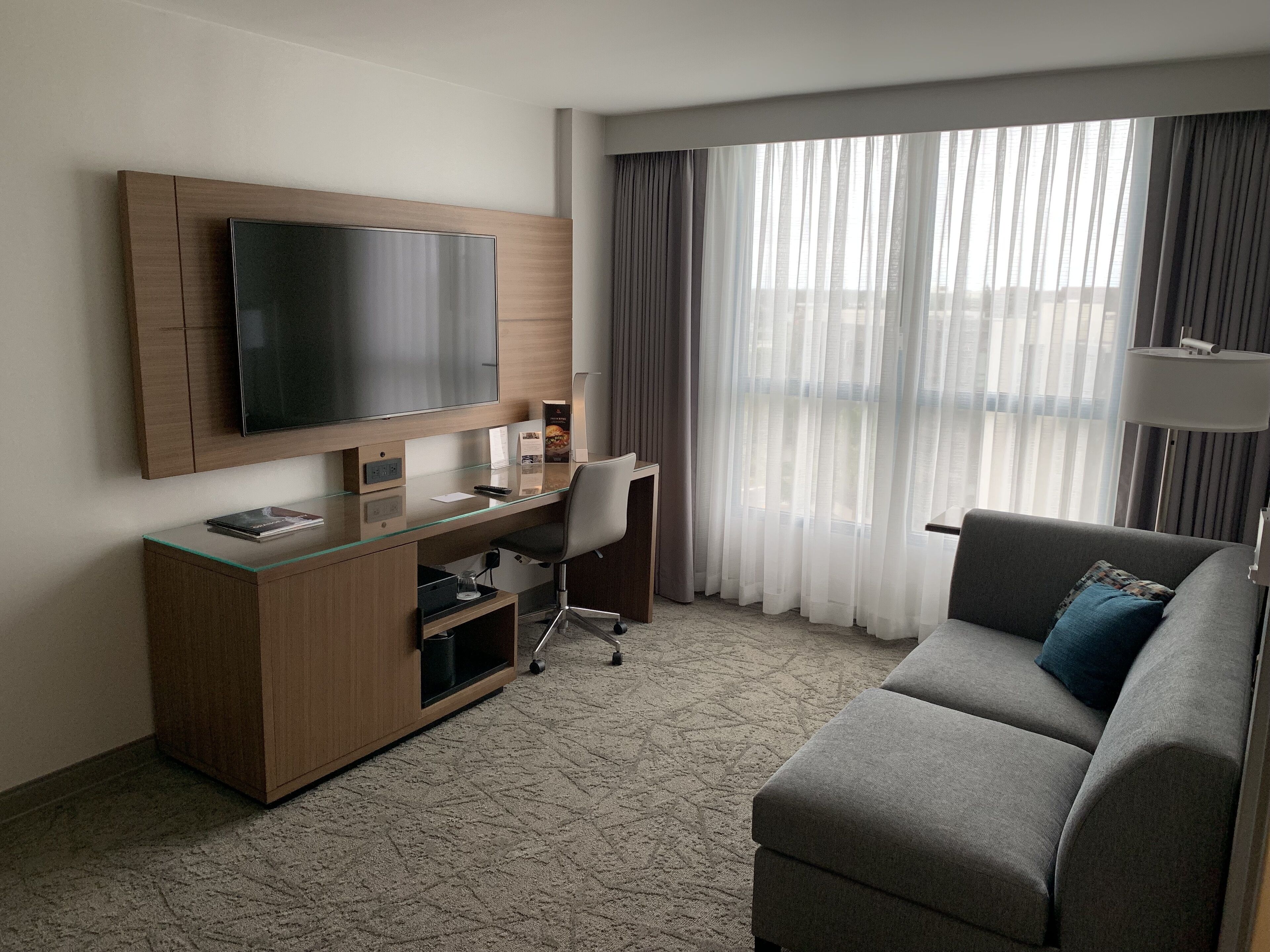 A modern hotel room with a flat-screen TV mounted on a wooden cabinet, a glass desk, a grey sofa, and a large window with sheer curtains.