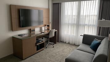 A modern hotel room with a flat-screen TV mounted on a wooden cabinet, a glass desk, a grey sofa, and a large window with sheer curtains.