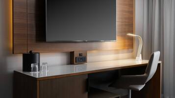 A modern hotel room with a large flat-screen TV mounted on the wall, a desk with a chair, a lamp, and a small table with glasses.