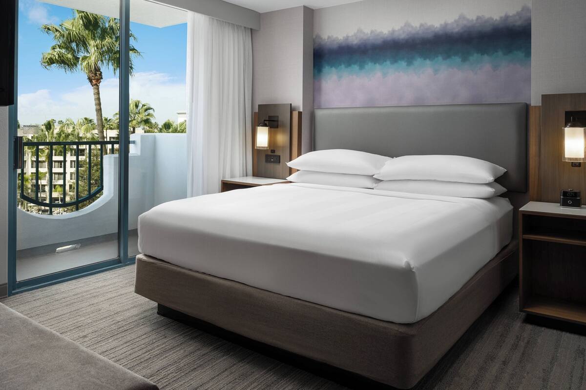 Habitación de hotel con una cama grande, vistas a palmeras, un balcón y un cuadro en la pared.