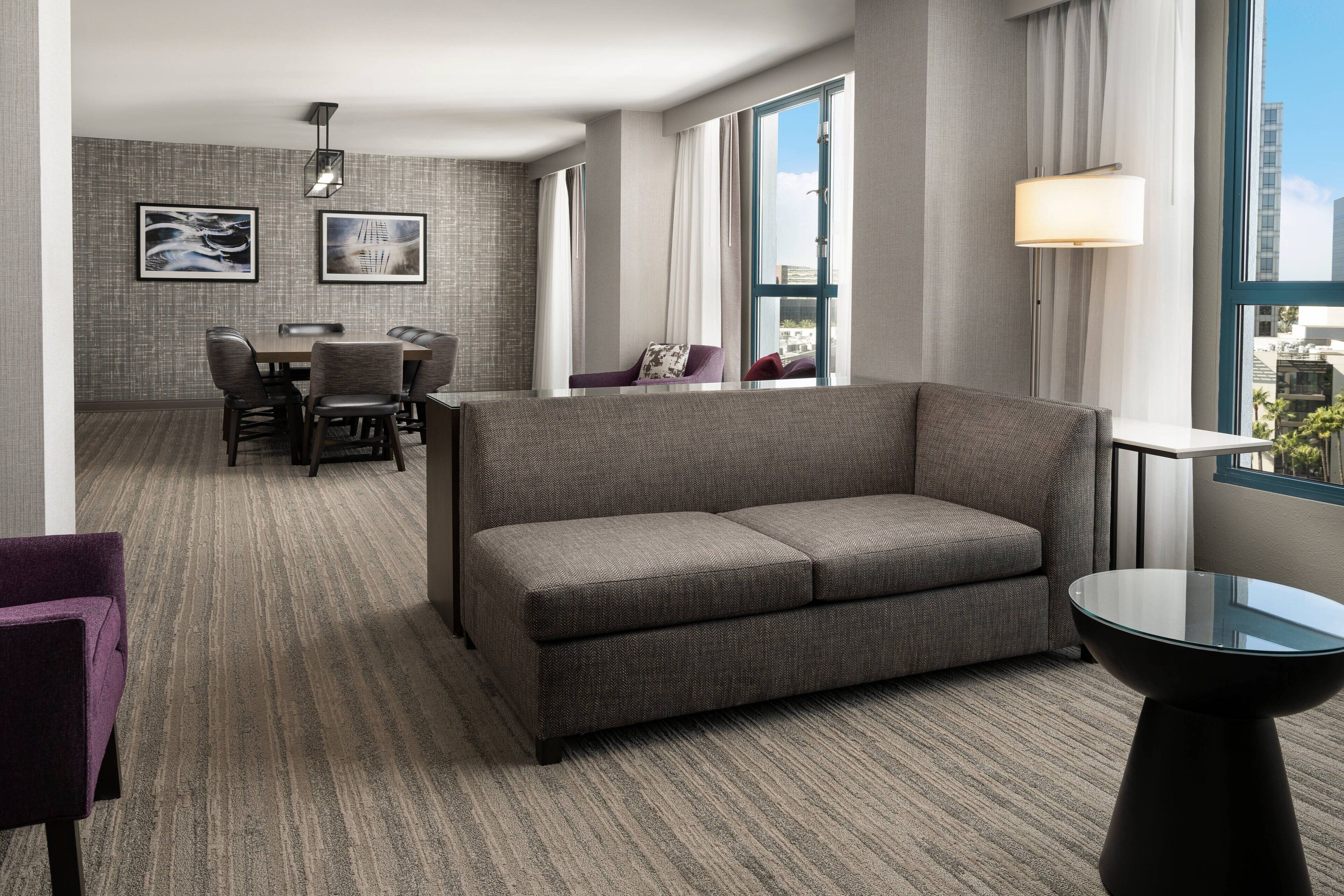 A modern hotel room with a grey sofa, a dining table with chairs, and a large window offering a city view.