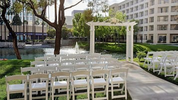 A wedding ceremony setup with white chairs and a white archway by a pond, surrounded by greenery and a building in the background.