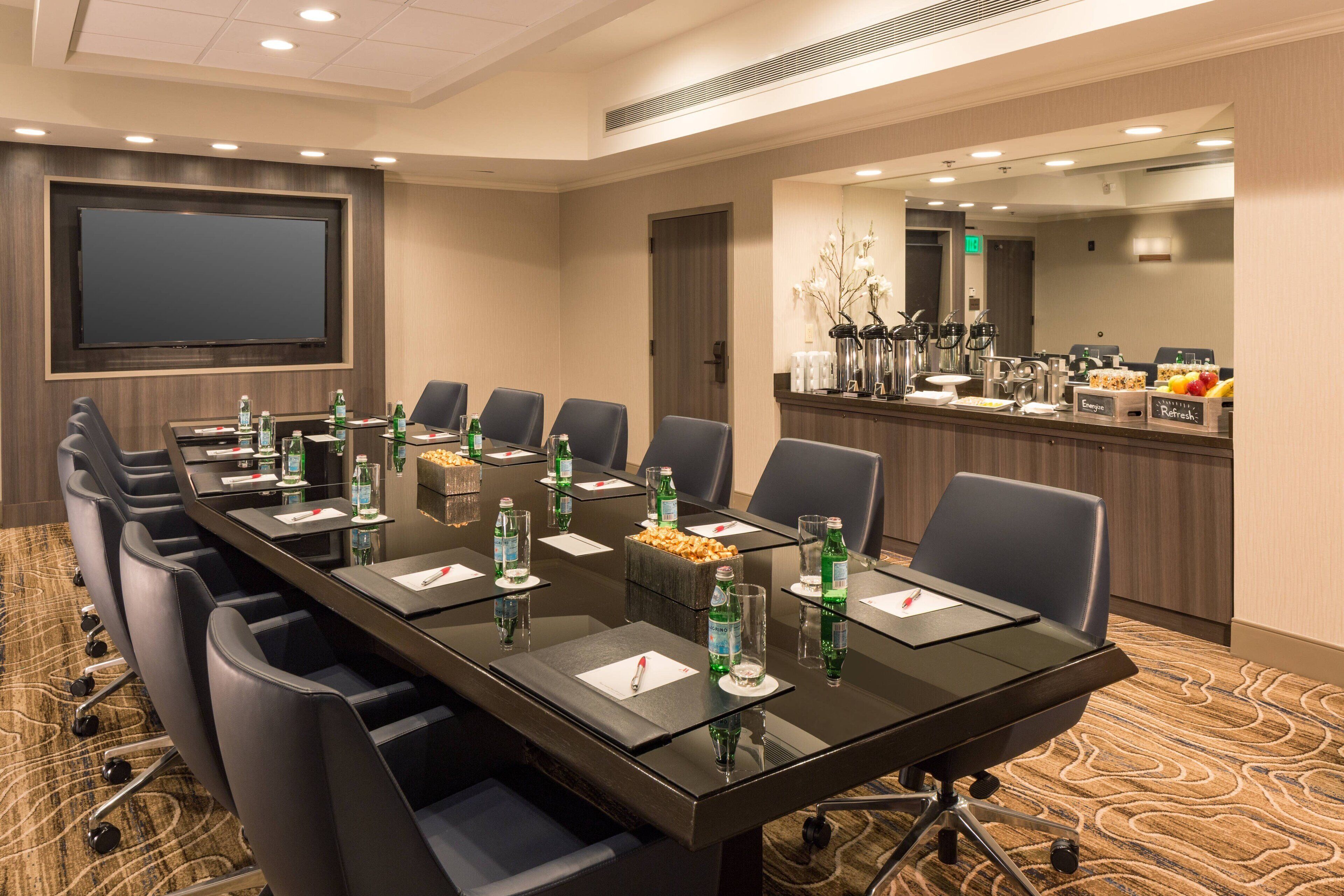 Un salón de conferencias con una mesa larga, sillas, una pantalla grande y una zona de bufé con comida y bebidas.
