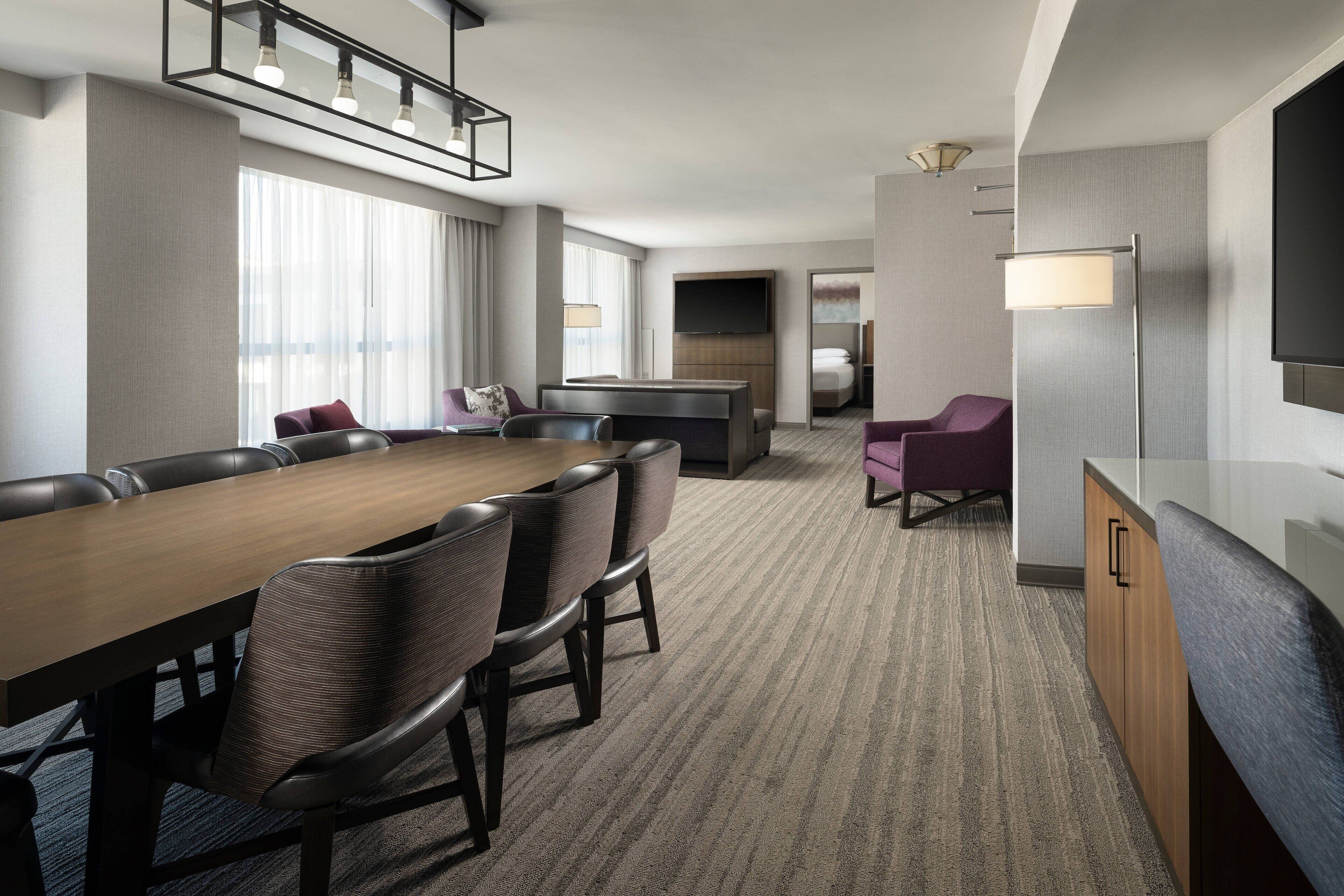 Habitación de hotel con una gran mesa de comedor, varias sillas, un sofá, un televisor y un minibar.