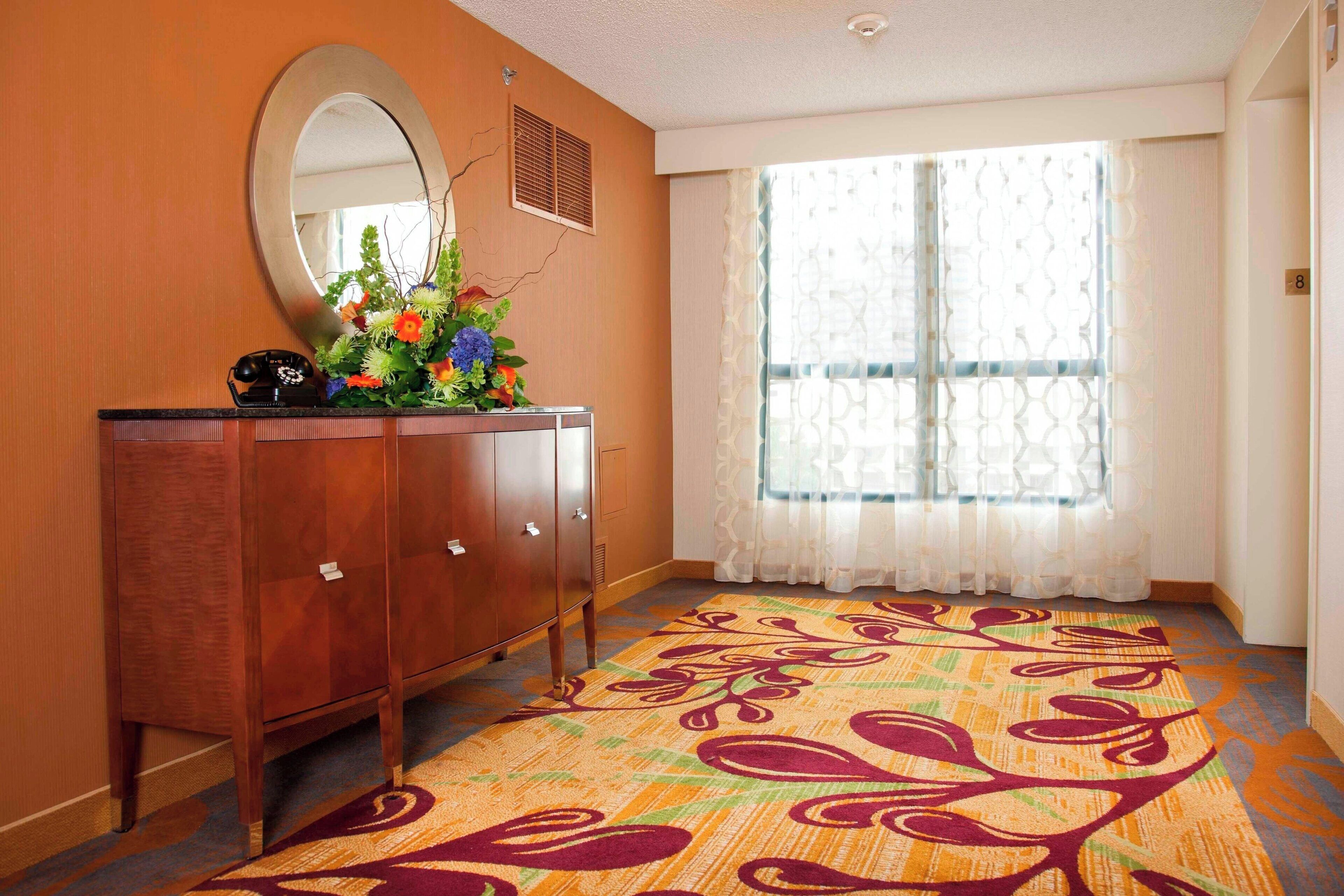 A hotel room with a wooden dresser, a mirror, a floral arrangement, and a patterned carpet.