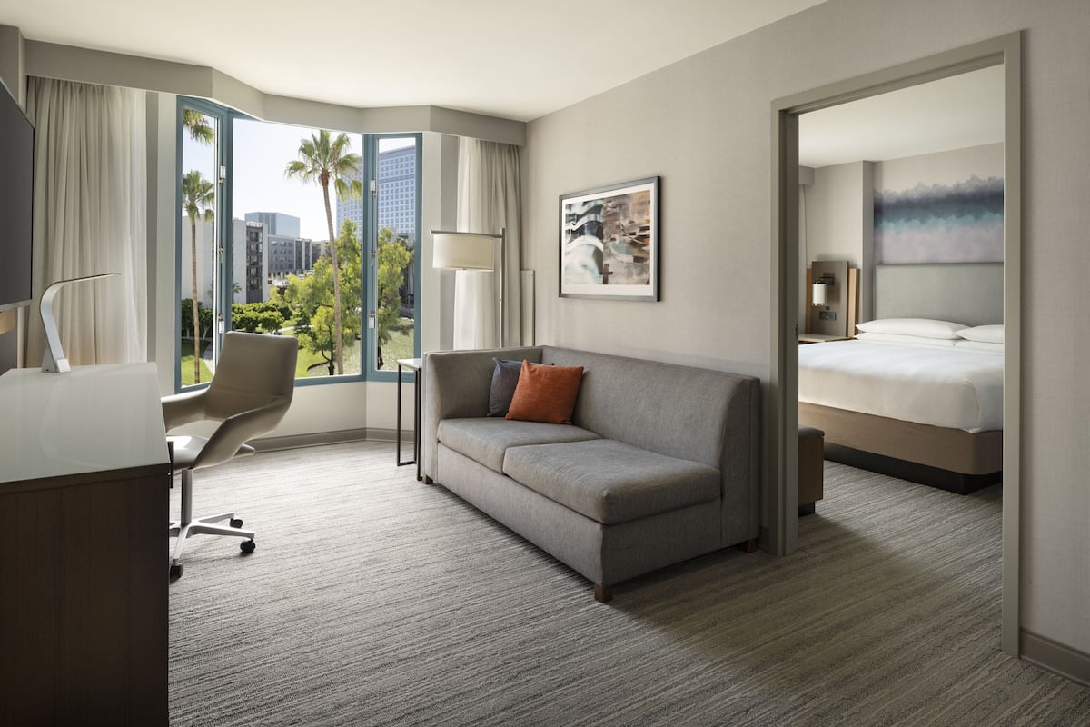 Habitación de hotel con sofá, escritorio, cama y ventana grande con vistas a palmeras y edificios.