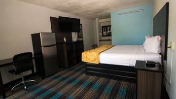 A hotel room with a bed, a desk, a chair, a refrigerator, a television, and a microwave.