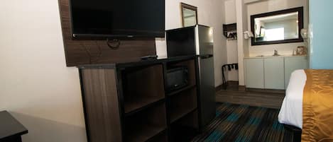 Una habitación de hotel con televisor de pantalla plana montado en la pared, una kitchenette pequeña con microondas y una cama con colcha estampada.