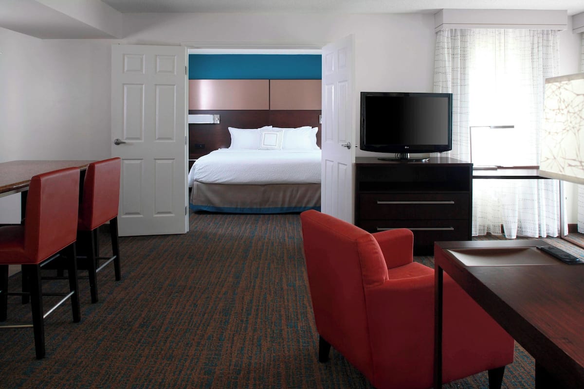A hotel room with a bed, a TV, a desk, and two red chairs.