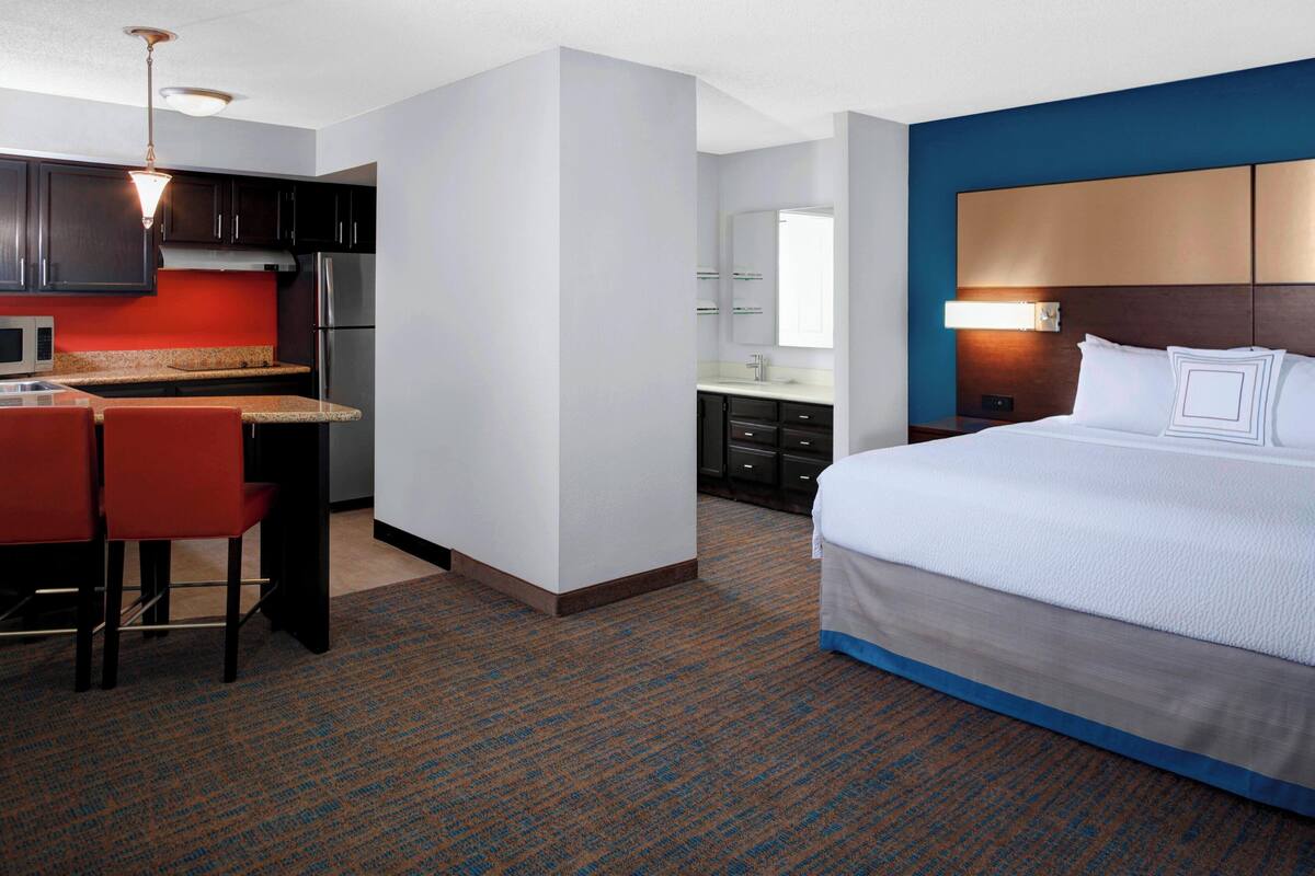 A hotel room with a bed, a desk with two chairs, a kitchenette, and a bathroom.