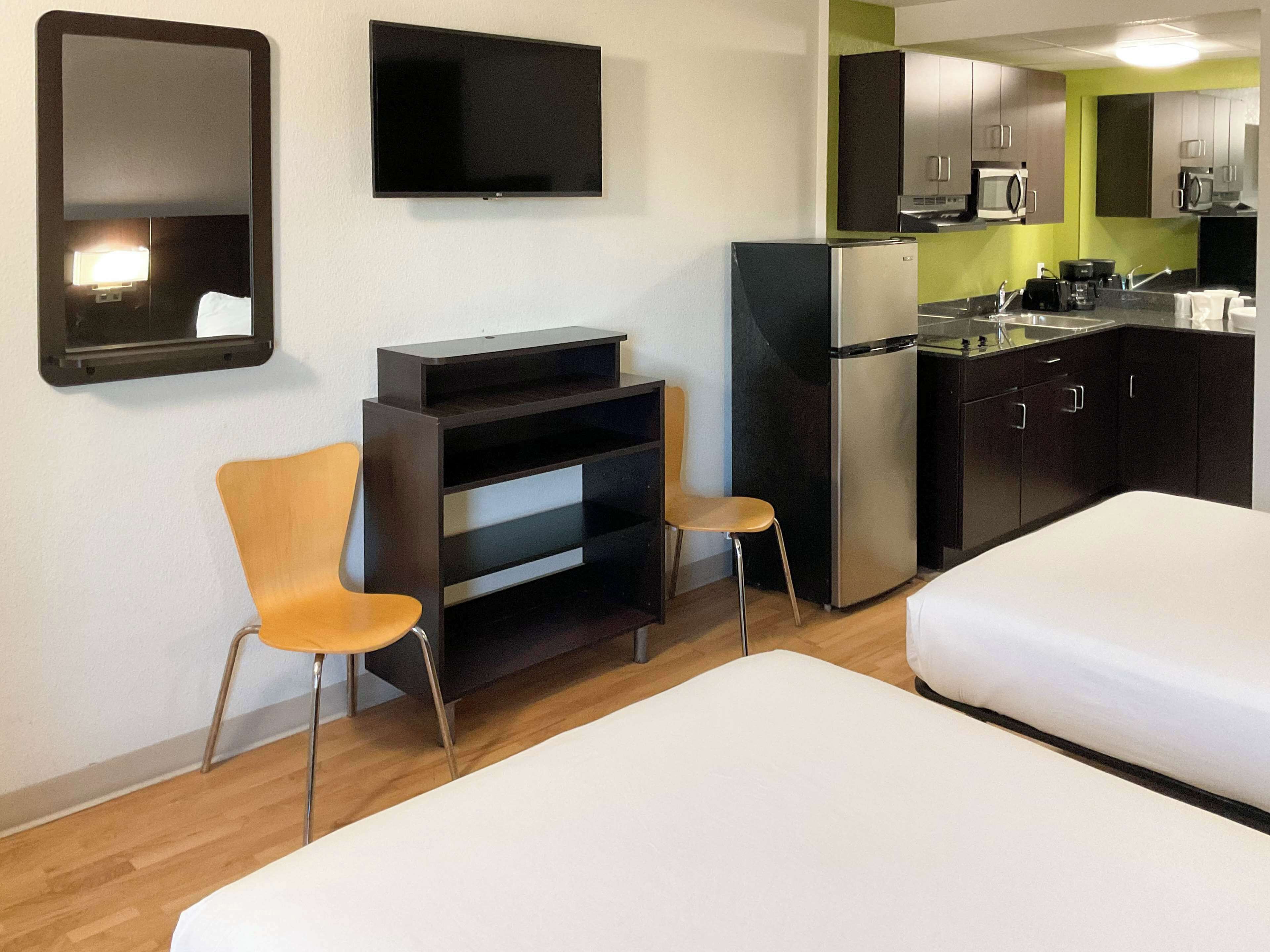 A compact hotel room with a kitchenette, two single beds, a TV, and a mirror.