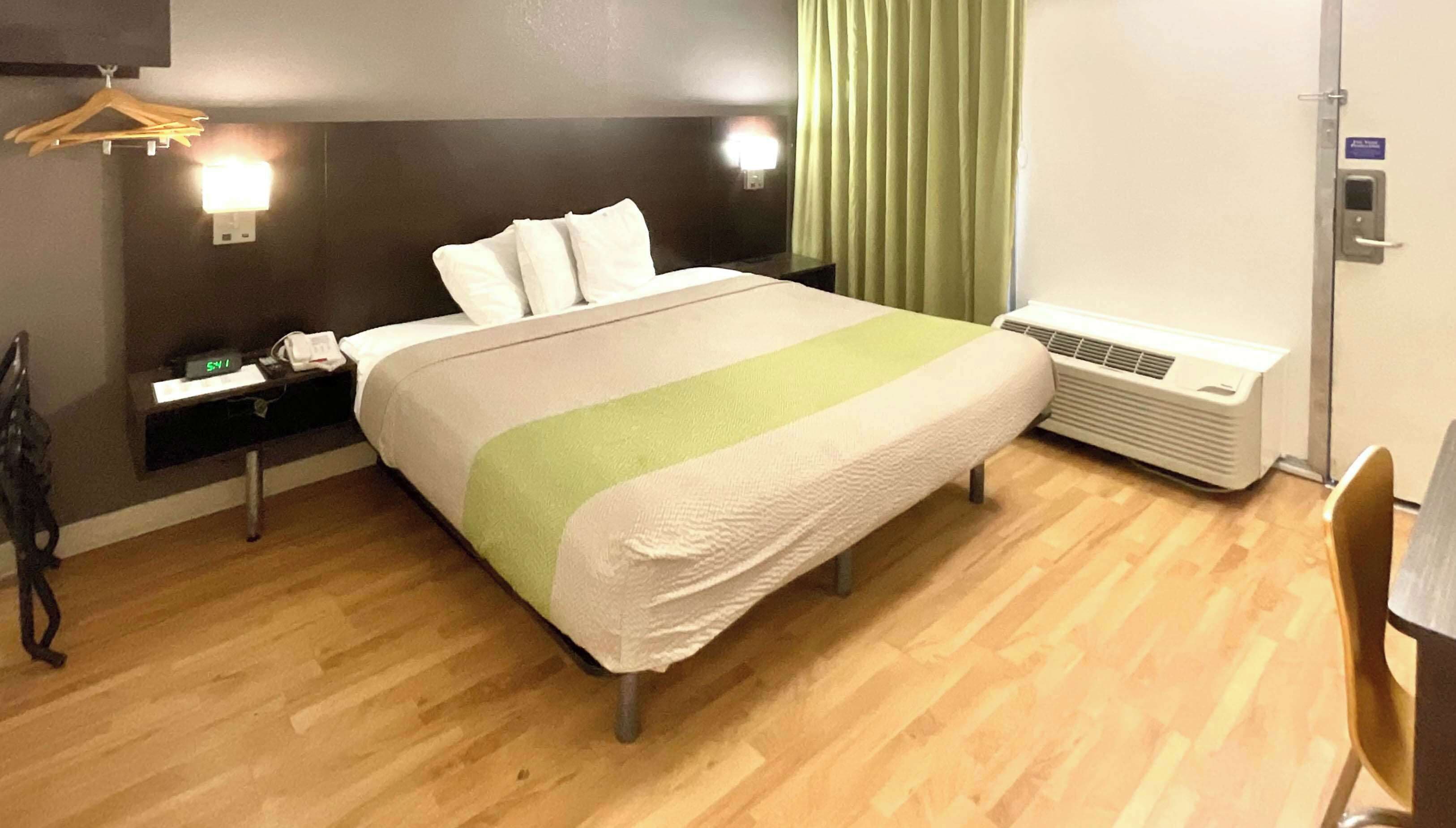 A hotel room with a bed, bedside table, lamp, air conditioner, and a chair.