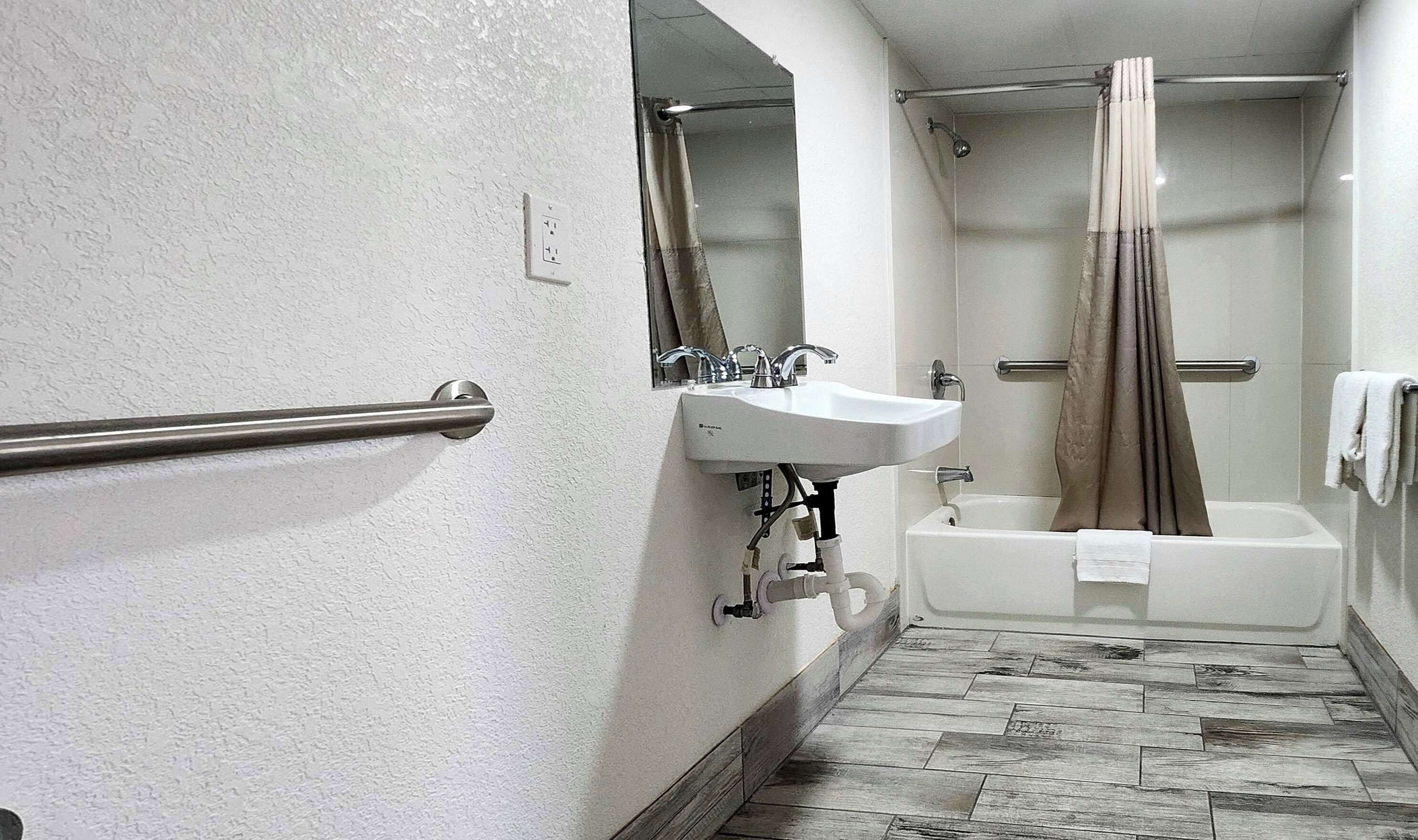 A narrow bathroom with a white sink, a mirror, a shower with a curtain, and a wooden floor.