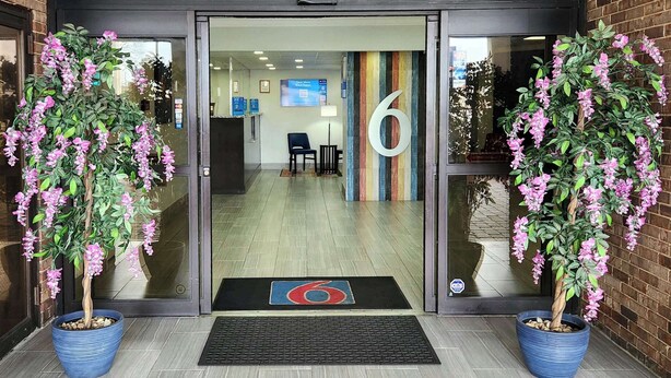 Entrada de hotel con puerta de vidrio, un cartel con el número 6 y dos macetas con flores rosas.