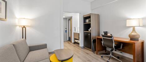 A modern hotel room with a sofa, a desk, a chair, a small yellow trash bin, and a lamp.