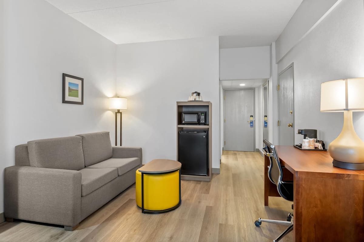 A modern hotel room with a sofa, a small round table, a desk with a lamp, a microwave, and a refrigerator.