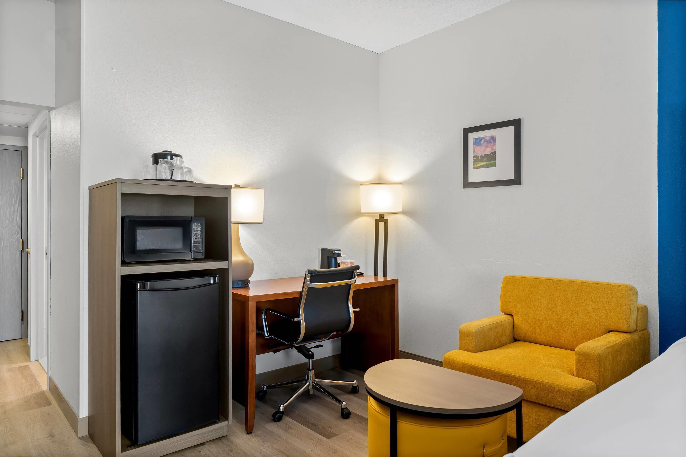 A compact hotel room with a kitchenette, a desk with two lamps, a yellow armchair, and a small table.