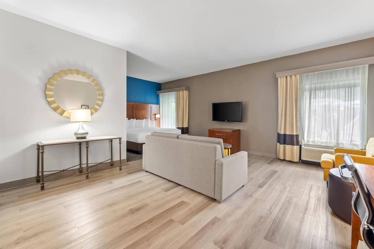 Ein Hotelzimmer mit Sofa, Fernseher, Nachttisch mit Lampe, Spiegel und einem fenster mit Vorhängen.