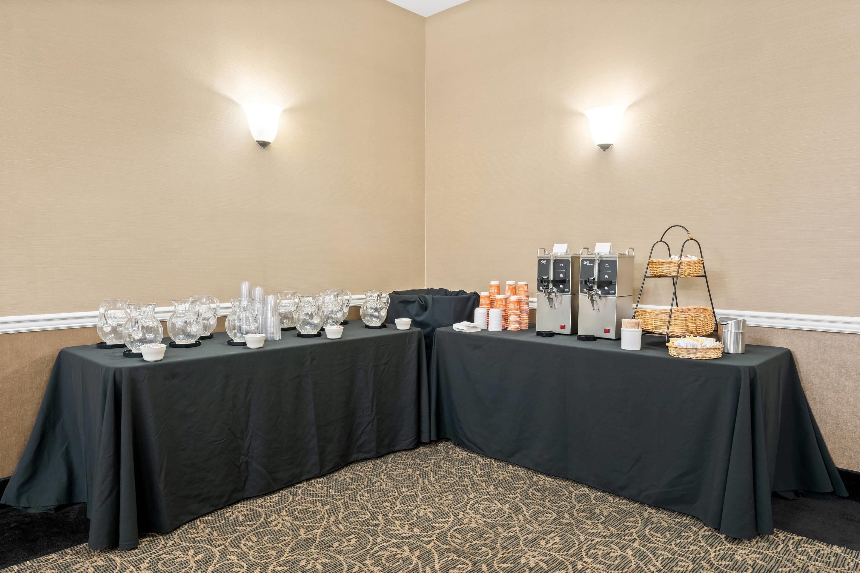 Ein Konferenzraum mit zwei Tischen, beide mit schwarzen Tischdecken bedeckt, ausgestattet mit Gläsern, Tassen und einer Kaffeemaschine.