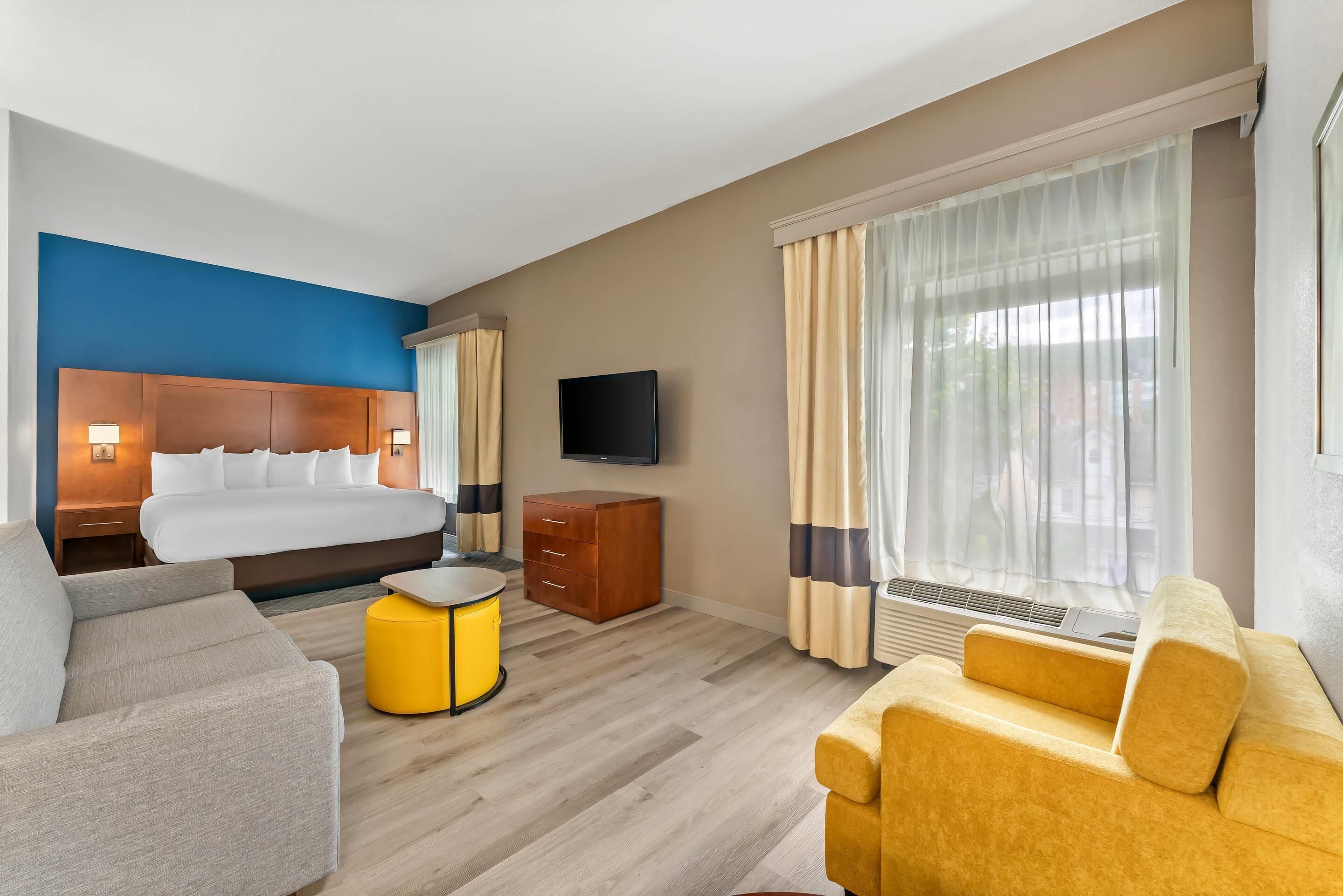 A hotel room with a bed, a TV, a sofa, a small table, and a window with curtains.