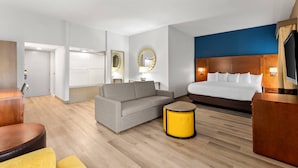 Una habitación de hotel con cama, sofá, una mesita auxiliar amarilla y otra de madera, además de un espejo.